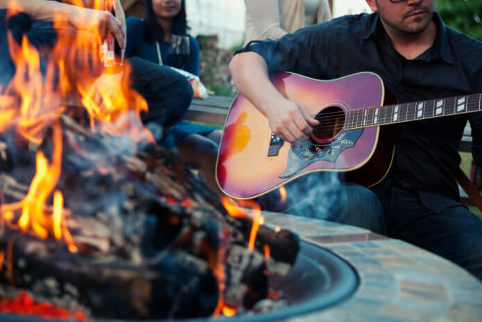 A man plays the guitar in the great outdoors as a campfire smolders next to him