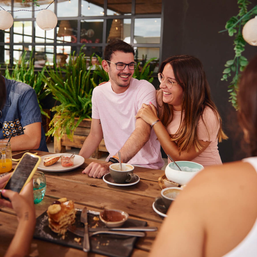 A young couple laughs while enjoying breakfast outside with friends