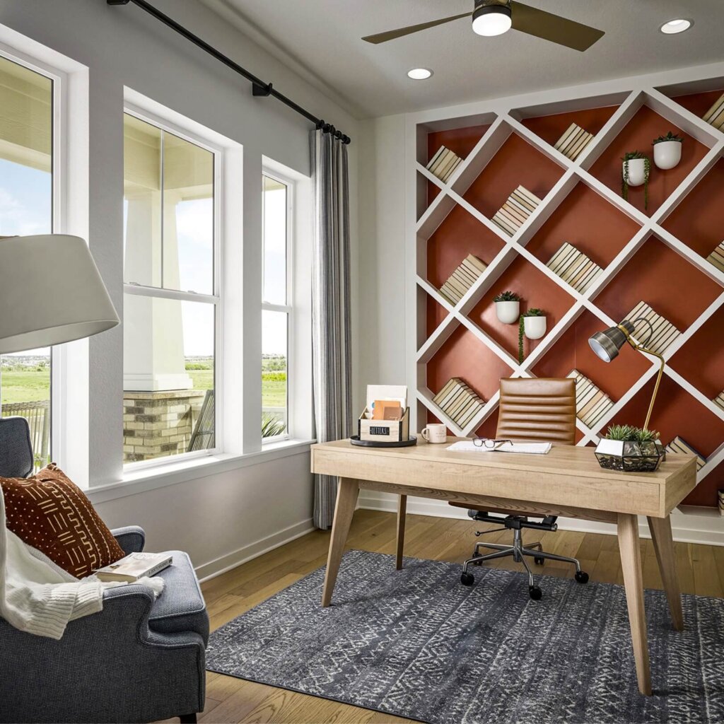 A modern home office in Kinston at Centerra features a wooden desk, beige chair, and geometric bookshelf filled with books. A large window lets in natural light, while a gray armchair and patterned rug complete this stylish model home ambiance in Loveland.