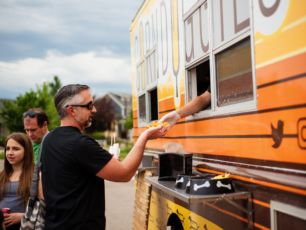 A man receives food from a person inside a brightly colored food truck while two people stand nearby, all enjoying the lively atmosphere at Centerra in Loveland, Colorado.