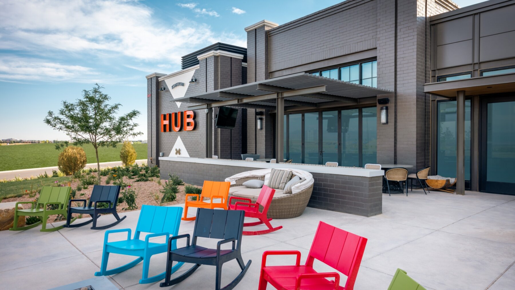 Outdoor seating area with colorful rocking chairs and modern building labeled "HUB" under a blue sky, perfect for savoring local flavors from Centerra restaurants.