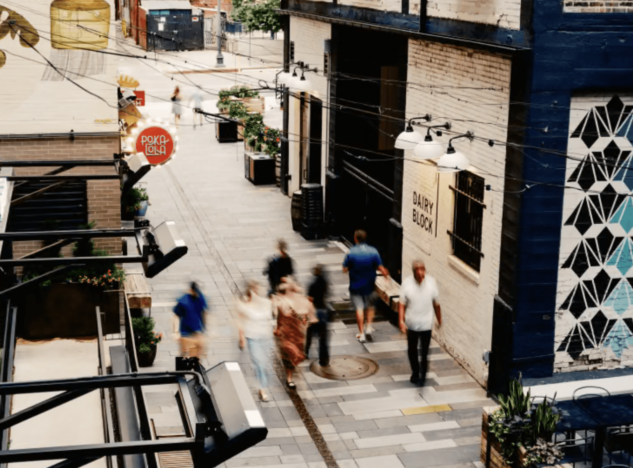 An urban alleyway in Dairy Block with several blurred people walking, outdoor string lights, wall art, and Realberry shop signs visible during daylight.