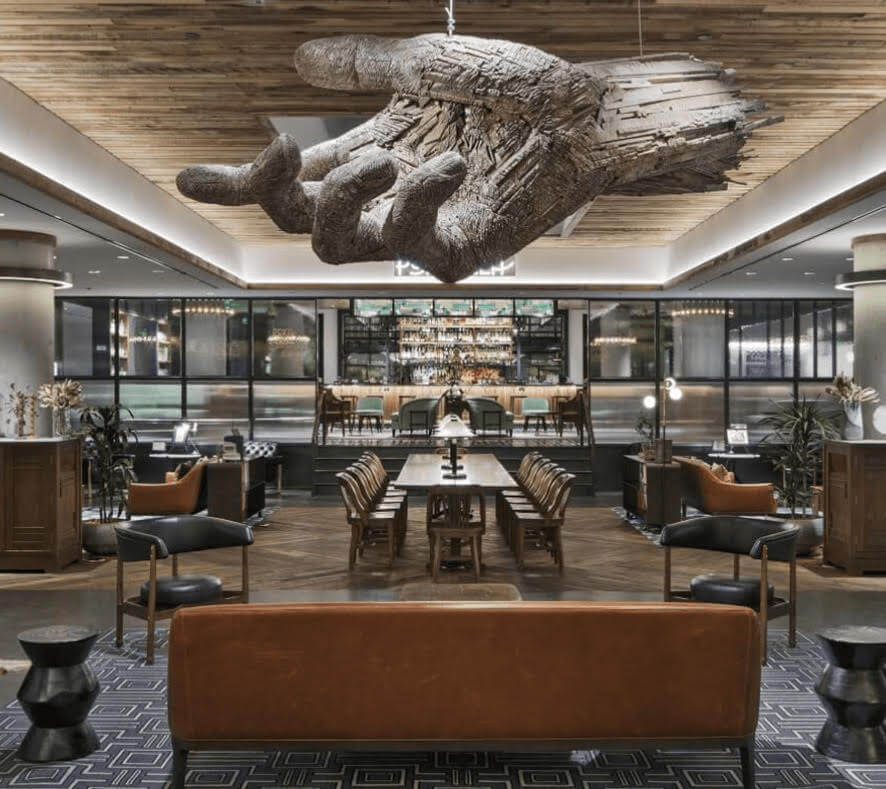 A modern hotel lounge at Dairy Block with leather seating, a central wooden table, and a striking wooden hand sculpture hanging from the ceiling.