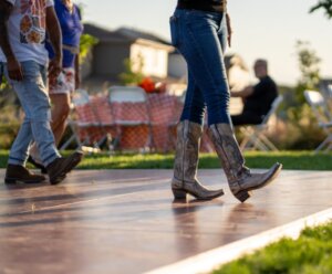 People wearing jeans and cowboy boots walk on an outdoor wooden platform; tables and chairs are set up in the background on the grass.