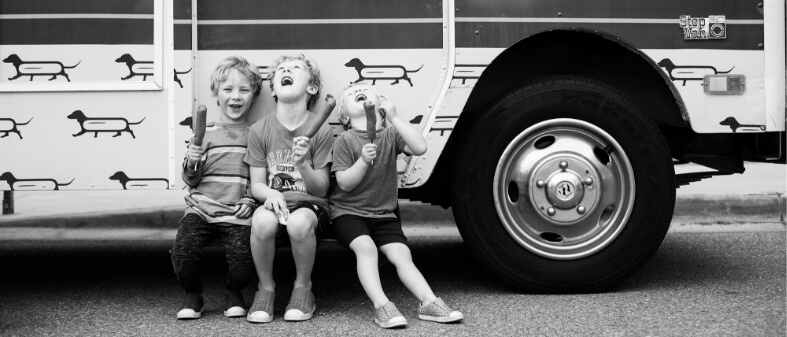 Black and white image of three kids eating corndogs while leaning against a food tuck