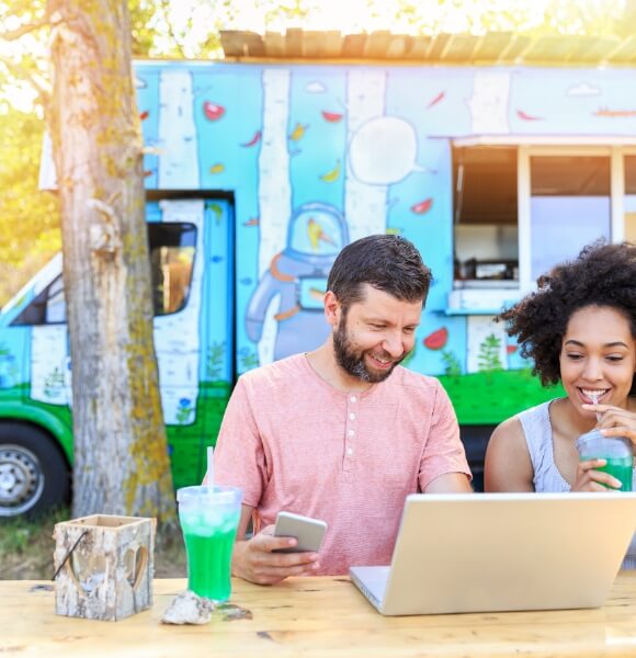 Image of couple using a computer on a picnic table with a food truck in the background