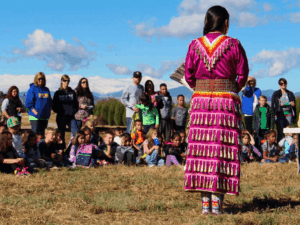 A woman in traditional attire stands outdoors at Centerra, facing a seated audience of children and adults on a grassy field under a blue sky, where families gather to explore new homes and enjoy the vibrant community atmosphere.