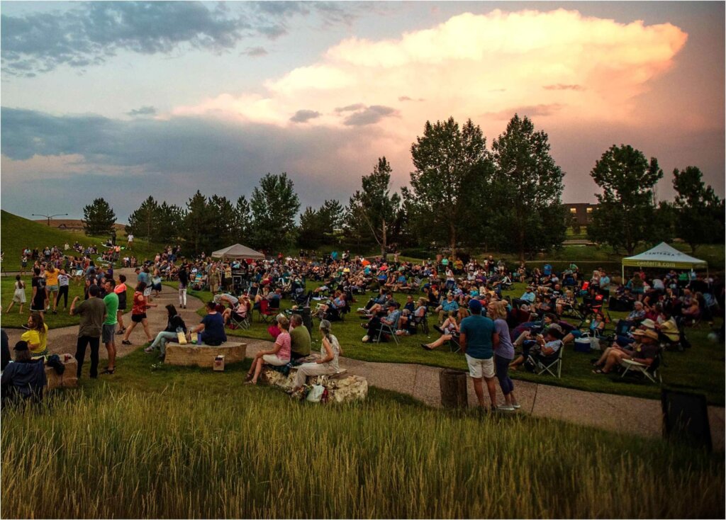 A crowd of people sitting in a grassy field at sunset, enjoying the Certified Wild beauty of nature.