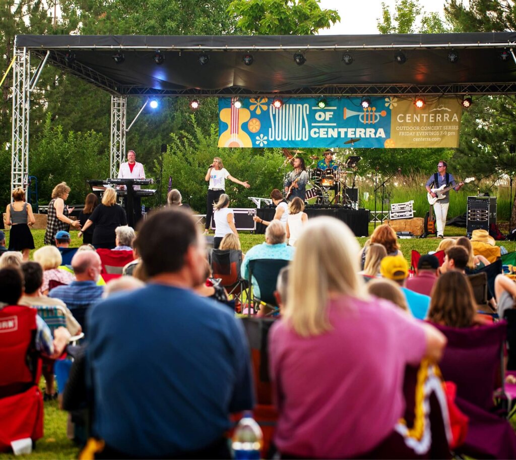 A band performs on an outdoor stage at the "Sounds of Centerra" concert in Loveland, Colorado, while a seated audience watches. Bright lights illuminate the stage, and trees are visible in the background. Nearby new homes add to the charm of this vibrant community event.