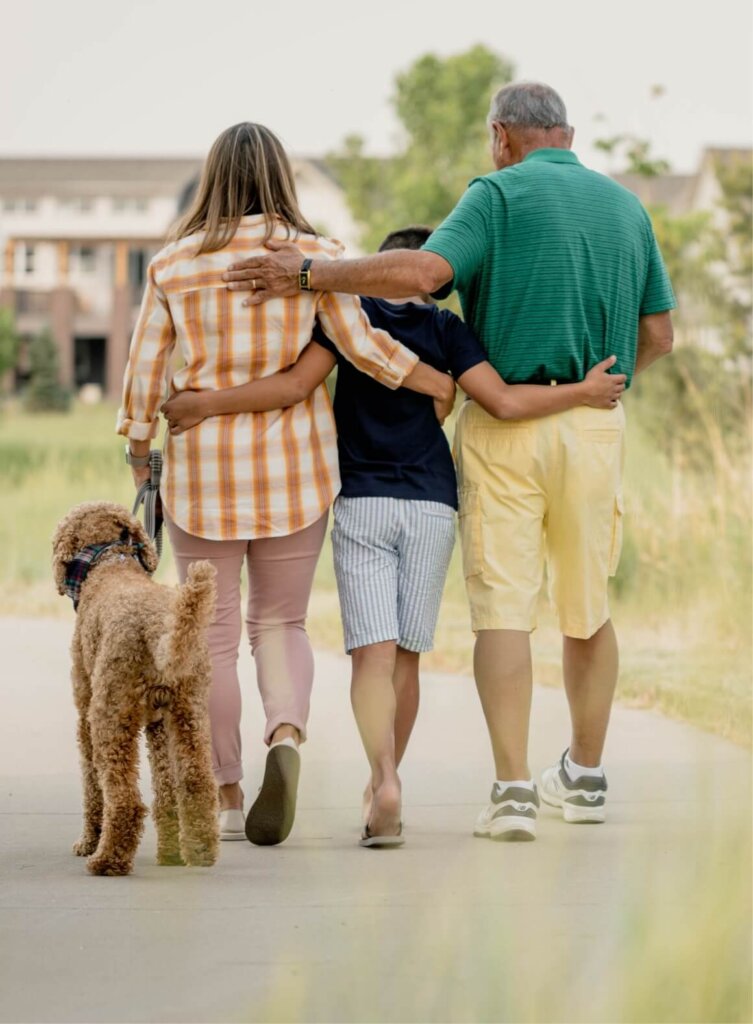 Three people walking on a path with their arms around each other, accompanied by a dog. In the background, you can see the greenery and charming new homes of Centerra in Loveland, Colorado.