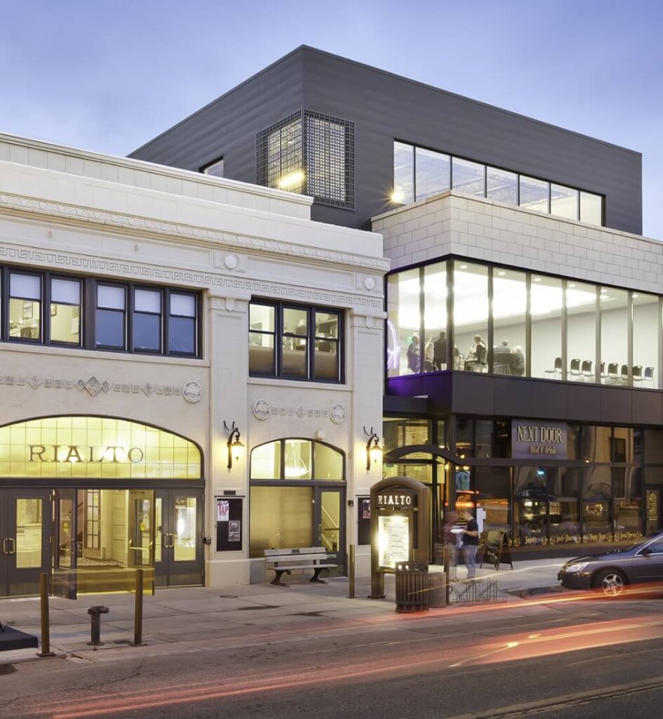 A street view of a modern building complex in Loveland, Colorado. The Rialto building is on the left with a vintage facade, while the newer, glass-fronted New Door coworking space on the right adds a contemporary touch. Nearby Centerra offers new homes that blend seamlessly with this evolving urban landscape.