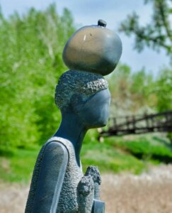 A bronze statue of a faceless figure with a round object balanced on its head, set against a backdrop of green trees and the partially visible bridge in Centerra, Loveland, Colorado—a serene spot near new homes.