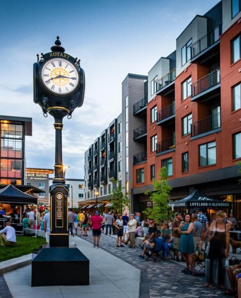 A tall clock in a busy outdoor scene at Centerra, surrounded by modern buildings and new homes, with numerous people walking and sitting, enjoying the evening in Loveland, Colorado.