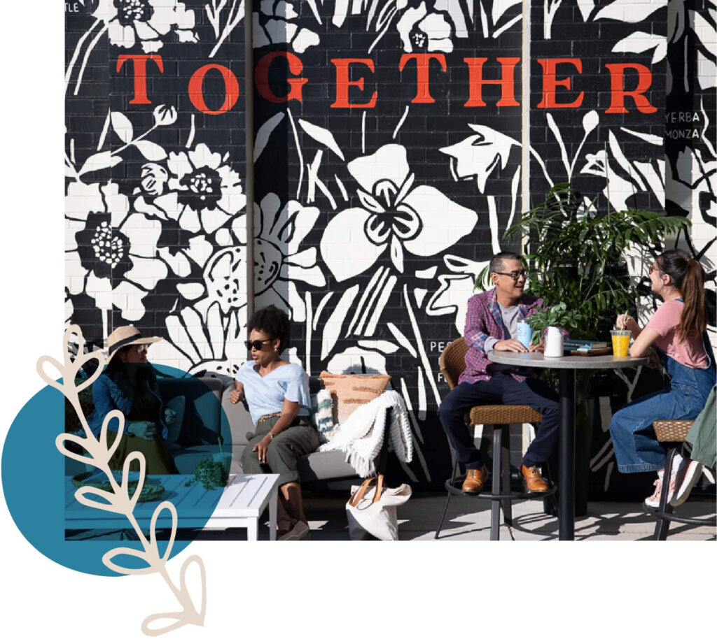Four people sit and stand around tables in an outdoor café in Centerra, Loveland, Colorado, with a large mural reading "TOGETHER" and floral designs on the wall behind them. There are drinks on the tables and a potted plant nearby, creating a perfect spot to discuss new homes in the area.