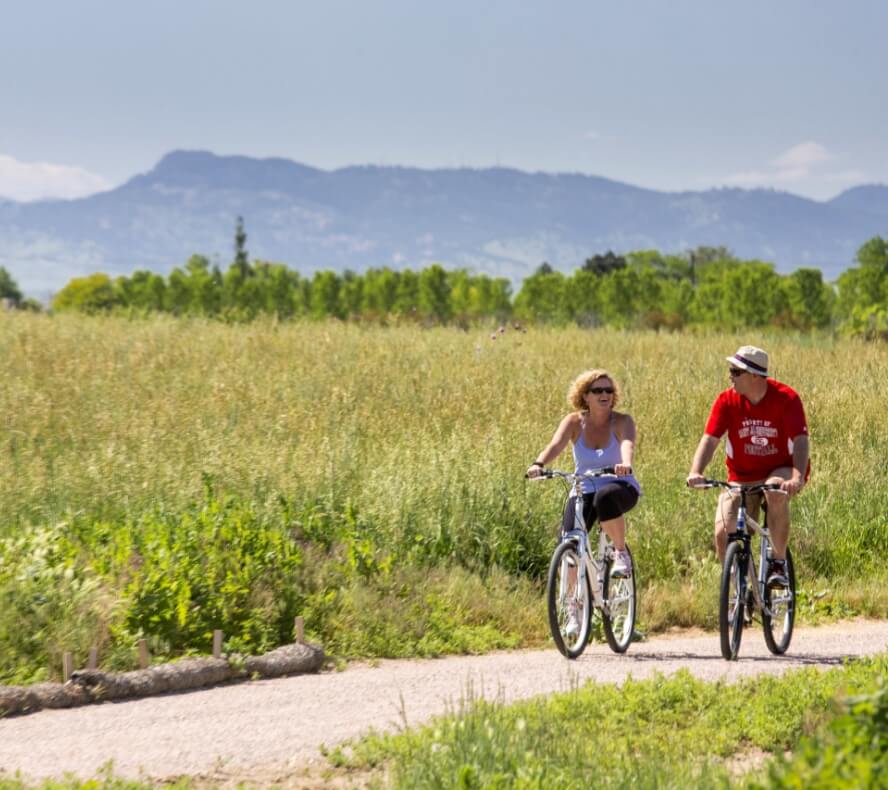 Two people ride bicycles on a paved path through a grassy field with mountains in the background on a clear day in Loveland, Colorado, near the vibrant community of Centerra.