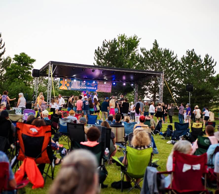 A crowd sitting on lawn chairs and standing in front of an outdoor stage enjoying a live musical performance in Centerra, Loveland Colorado, where new homes offer the perfect community vibe.