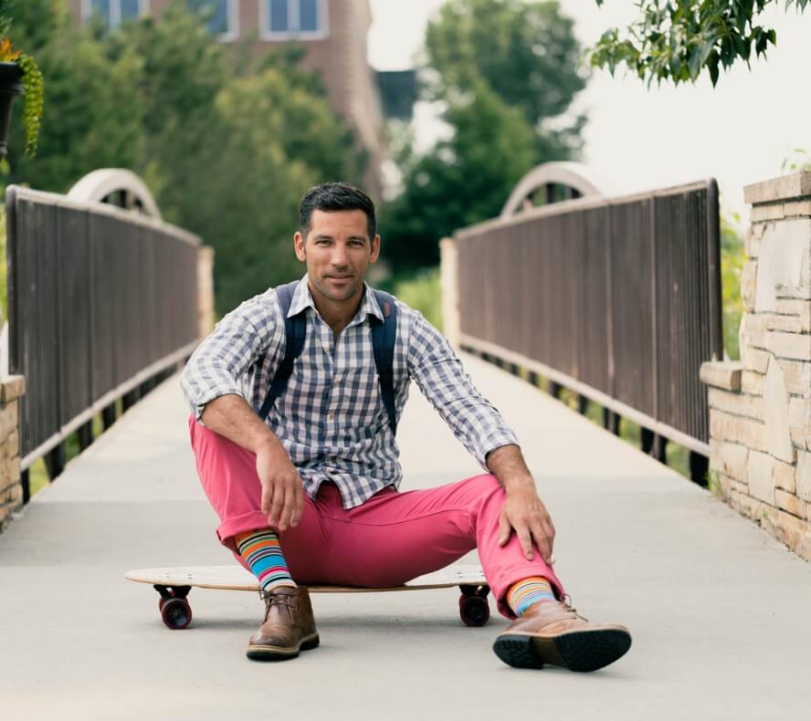 A man in pink pants and a checkered shirt sits on a skateboard on a bridge in Loveland, Colorado, smiling at the camera. He is wearing colorful striped socks and has a backpack. The backdrop captures the charm of Centerra's new homes.