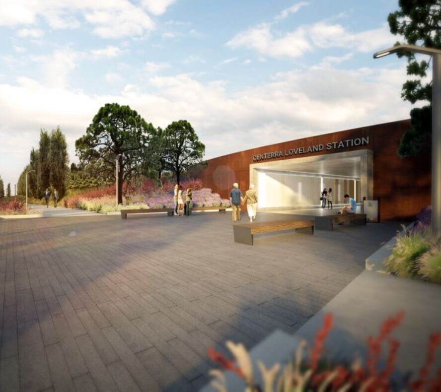 A modern train station marked "Centerra Loveland Station" with people standing and sitting outside, surrounded by trees and landscaping in Loveland, Colorado. The sky is partly cloudy.
