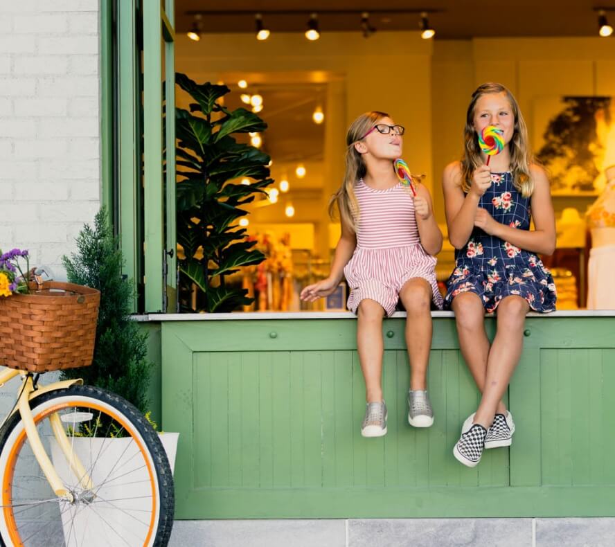 Two children sit on a windowsill in their new home, enjoying lollipops. One wears a striped dress and glasses, the other a floral dress. Nearby, a bicycle with a wicker basket is parked. The background reveals the lit and decorated interior of their cozy Centerra residence in Loveland, Colorado.