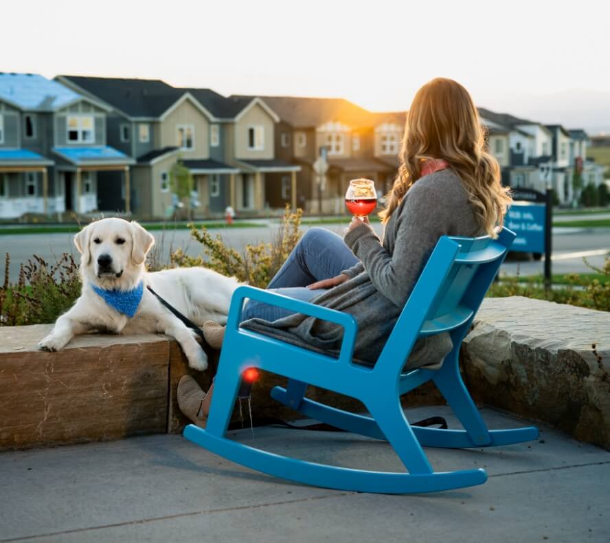 A person sits in a blue rocking chair holding a drink while a dog rests nearby. They are outdoors in Loveland, Colorado, facing a row of new homes at sunset.