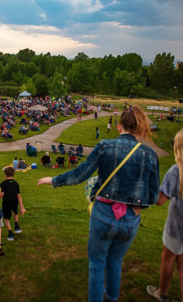 A person in a denim jacket and a crossbody bag walks down a grassy hill toward a large outdoor gathering with people sitting on chairs and blankets, savoring the vibrant community spirit that Centerra in Loveland, Colorado offers.