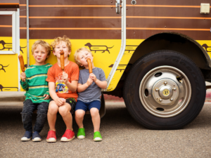Three young children sitting on the bumper of a food truck in Centerra, Loveland, Colorado, each holding and eating a corn dog.
