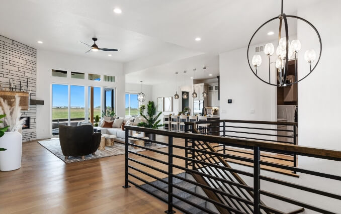 Open-concept living area with modern furnishings, a large chandelier, and a staircase leading down. The room has large windows with a view of the grassy landscape typical of Loveland, Colorado, and a kitchen is visible in the background. Perfect for those seeking new homes in Centerra.