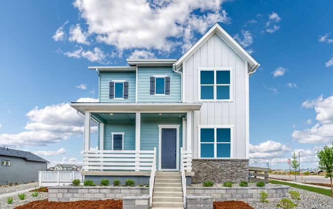 A two-story light blue house with white trim, a front porch, and a well-manicured yard in Loveland Colorado's Centerra neighborhood. Clear blue sky with white clouds in the background.