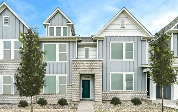 A modern two-story townhouse with a light brick and gray exterior, large windows, and a teal front door. Located in Centerra, Loveland Colorado, it is flanked by two small trees and has a stone path leading to the entrance—an ideal choice among new homes in the area.
