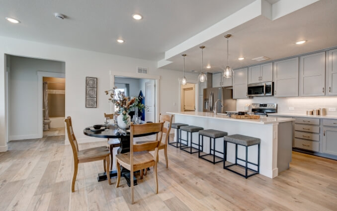 Modern kitchen and dining area in a new home at Centerra, Loveland, Colorado. Features light wood flooring, white countertops, stainless steel appliances, pendant lights, and a round table with wooden chairs. The kitchen has gray cabinets and barstools.