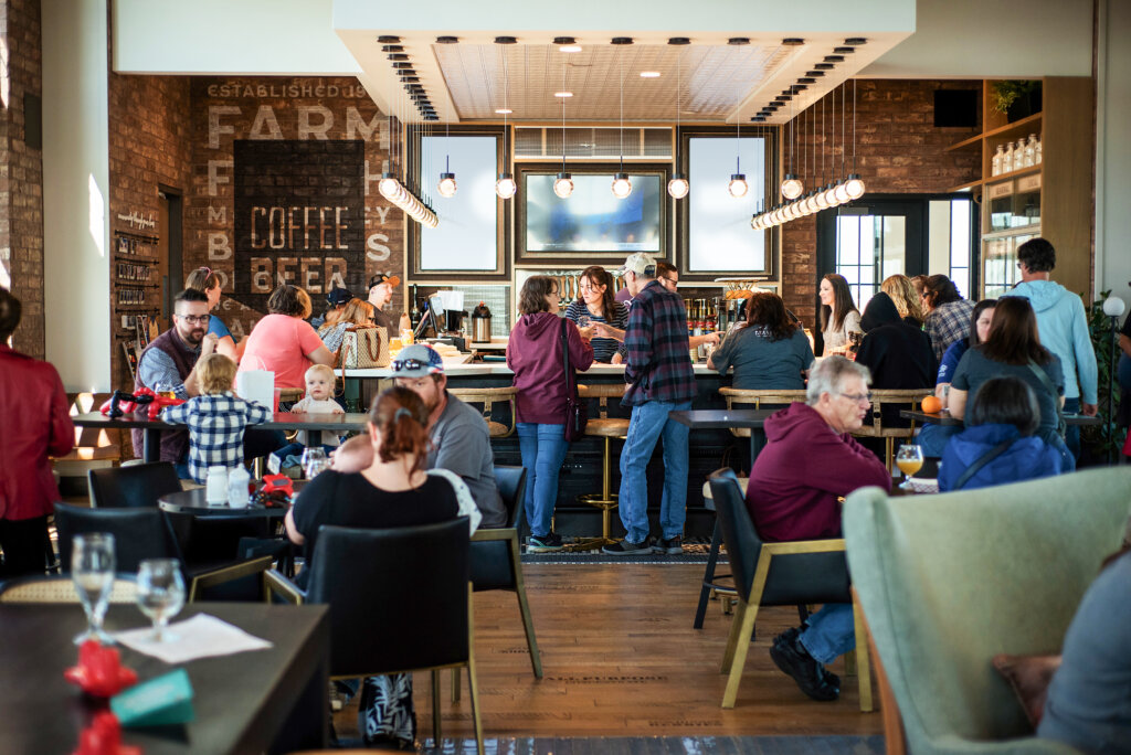 People are gathered inside a modern café or bar, sitting and standing around tables and the counter, with drinks and food visible on tables.