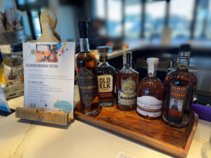 Five bottles of Colorado bourbon are displayed on a wooden tray next to a sign promoting a Colorado bourbon tasting event at a bar counter in Centerra, Loveland Colorado.