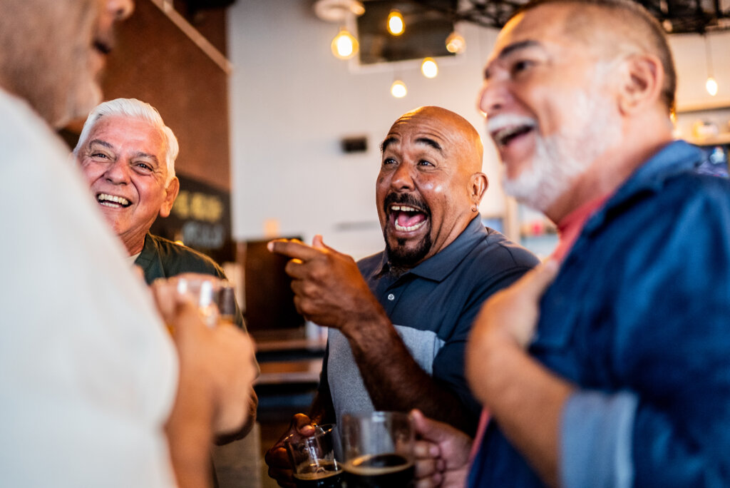 Four older men standing close together, laughing and holding drinks in a well-lit indoor setting with hanging lights in the background.