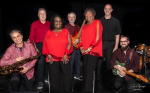 Seven musicians pose together on stage with instruments, wearing coordinated red and black outfits. Performing in Centerra, Loveland Colorado, the group includes men and women, with a saxophone and guitar visible—just steps from stunning new homes.