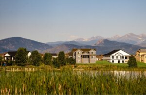 Modern houses line a residential neighborhood in Centerra, Loveland Colorado, with mountains in the background and a pond with tall reeds in the foreground under a clear sky.