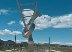 A large, metallic art sculpture resembling a twisted ladder is installed at a street intersection in Loveland, with houses and mountains in the background under a blue sky.