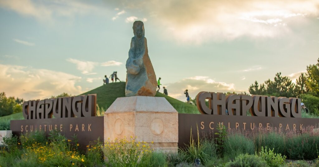 Entrance to Chapungu Sculpture Park in Centerra, Loveland, Colorado, featuring a large stone sculpture, metal signs, and landscaped greenery; people can be seen on a grassy hill in the background.