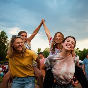 Two women give piggyback rides to two smiling girls, with one pair holding hands in the air. They're outdoors at Kinston Centerra, surrounded by trees and a cloudy sky, capturing the joy of community spirit near the model homes Loveland has to offer.