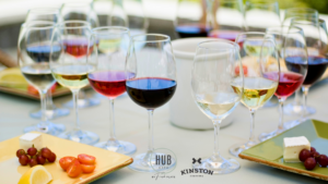 A table set with wine glasses containing red, white, and rosé wines sits ready for guests, accompanied by plates of cheese, grapes, and cherry tomatoes. Hub Cafe and Kinston logos are visible—the perfect setting to unwind after touring new homes in Loveland Colorado.
