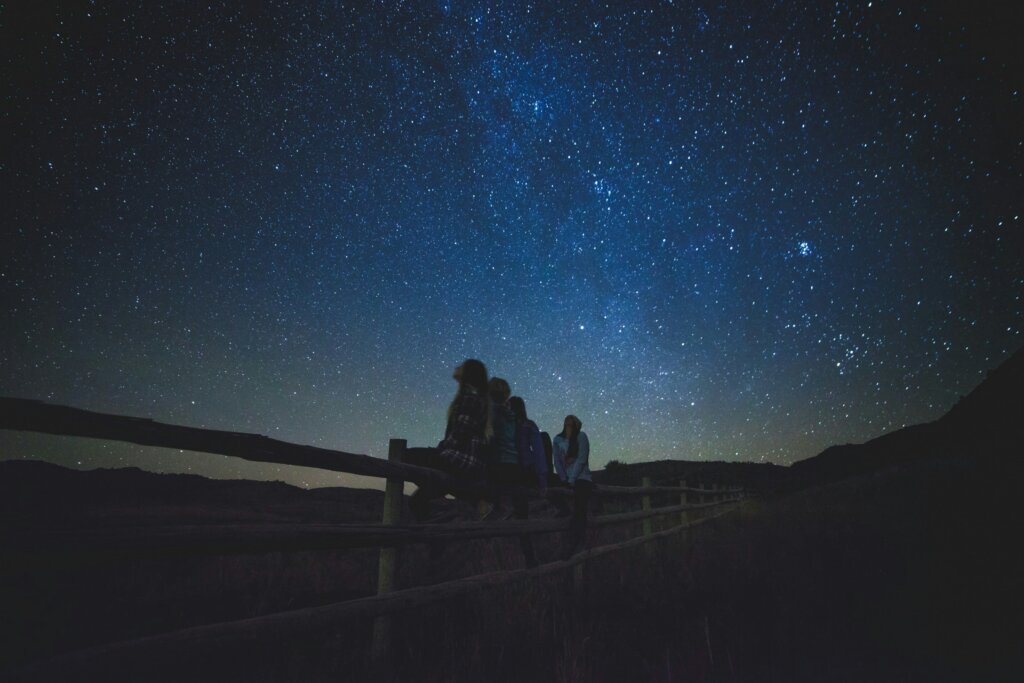 Five people sit on a wooden fence at night, silhouetted against a clear sky filled with visible stars and a faint Milky Way, creating a serene stargazing event shared by the group.