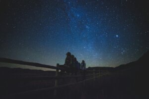 Five people sit on a wooden fence at night, silhouetted against a clear sky filled with visible stars and a faint Milky Way, creating a serene stargazing event shared by the group.