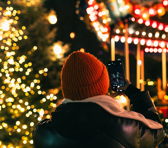 A person in a red knit hat takes a photo with a smartphone near a lit Christmas tree and a brightly illuminated carousel at night in Centerra, capturing the festive spirit of Loveland, Colorado’s vibrant new homes community.