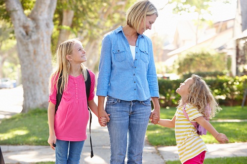 Mother And Daughters Walking To School Holding Hands On Suburban Street
